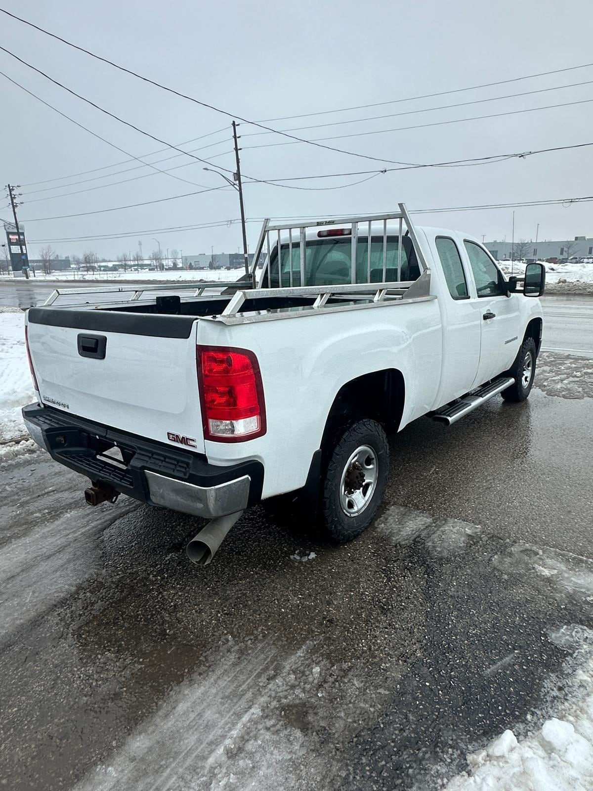 2007 GMC 2500HD DURAMAX DIESEL SIERRA 2500HD 6.6 DURMAX DIESEL WT EXTENDED CAB STD White rear three-quarter exterior view showing truck bed with rack and red taillights in snowy lot.
